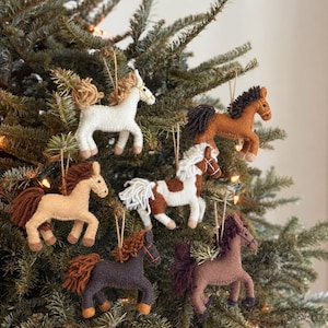 May include: A collection of six felt horse ornaments hanging from tree branches. The horses are crafted in various colors including white, tan, brown, dark brown, and a white horse with brown patches, each featuring yarn manes and tails.