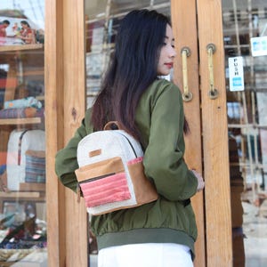 Peut inclure: Un petit sac à dos composé d'un corps principal en tissu tissé crème, de panneaux latéraux en suédine beige et d'une poche avant en tissu texturé à motifs rouges et blancs. Le sac présente une petite étiquette rectangulaire marron sur le haut.