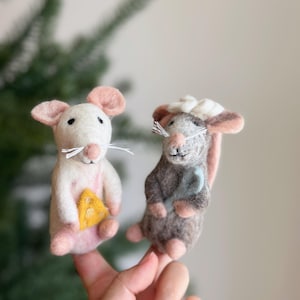 May include: Two felt mouse figurines. One is white with pink ears and feet, holding a yellow cheese wedge. The other is grey and brown, wearing a white hat and holding a blue object. Both have white whiskers.