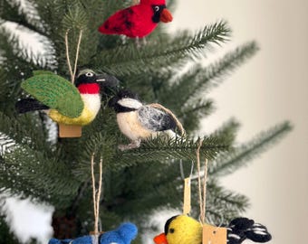 May include: Five felt bird decorations hanging on a green Christmas tree. The birds are a cardinal, a hummingbird, a chickadee, a blue jay, and a goldfinch.