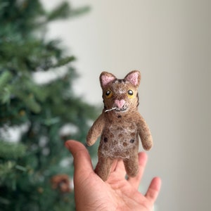 May include: A small, brown and black felted cat with pink ears and yellow eyes. The cat is holding a small white string in its mouth.