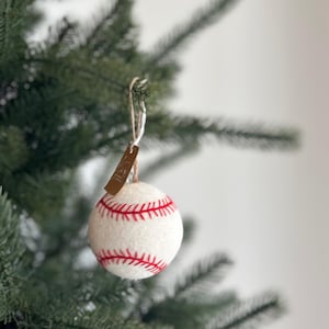 May include: A baseball-themed Christmas ornament. The ornament is white with red stitching, resembling a baseball. It hangs from a twine string and a small brown tag. The ornament is displayed on a green Christmas tree.