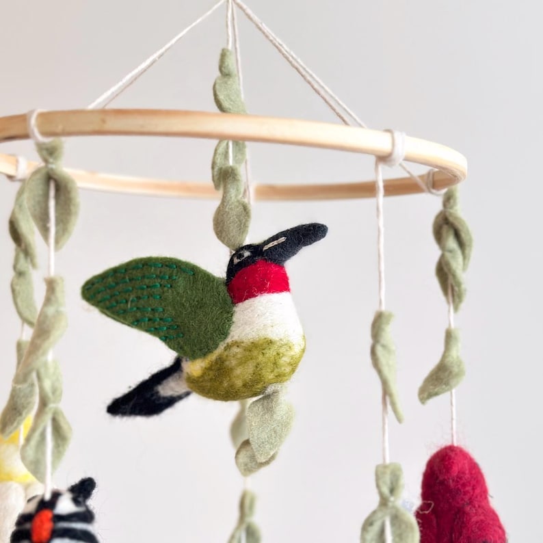 Puede incluir: Un m&oacute;vil para beb&eacute; de fieltro con un colibr&iacute; verde, rojo y blanco, hojas de fieltro verde y un anillo de madera.