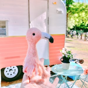 May include: A pink felt flamingo toy with black eyes and a black beak. The flamingo is being held in a person's hand in front of a pink and white vintage camper.
