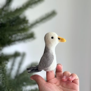 Peut inclure: Un doigt de marionnette en feutre représentant une mouette grise et blanche avec un bec jaune. La marionnette est perchée sur un doigt.
