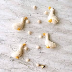 Puede incluir: Una guirnalda decorativa con cuatro gansos de fieltro blanco, cada uno con picos y patas amarillas, intercalados con pequeñas bolas de fieltro blanco. Los gansos están ensartados en un hilo blanco, creando un aspecto artesanal. También hay una pequeña campana dorada.