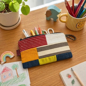 May include: A colorful, rectangular zippered pouch with a patchwork design in red, yellow, blue, and neutral tones. The pouch is filled with pens and pencils. A small ceramic rainbow and a drawing of a house are also visible.