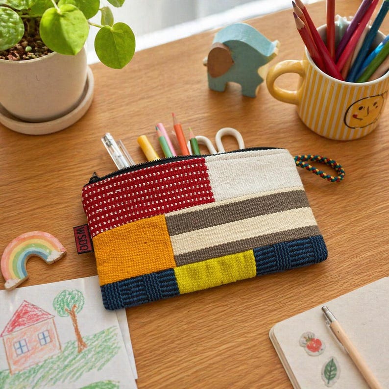 May include: A multi-coloured rectangular zipped pouch with a patchwork design in red, yellow, blue, and neutral tones. The pouch is filled with pens and pencils. A small ceramic rainbow and a drawing of a house are also visible.