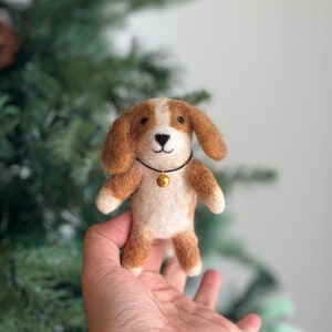 May include: A small, brown and white felt dog ornament with a gold bell around its neck. The ornament is being held in a person's hand.