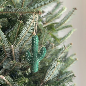 May include: A green felt cactus Christmas ornament with white stitching, hanging from a natural twine string. The ornament is displayed on a green Christmas tree, creating a festive holiday scene.