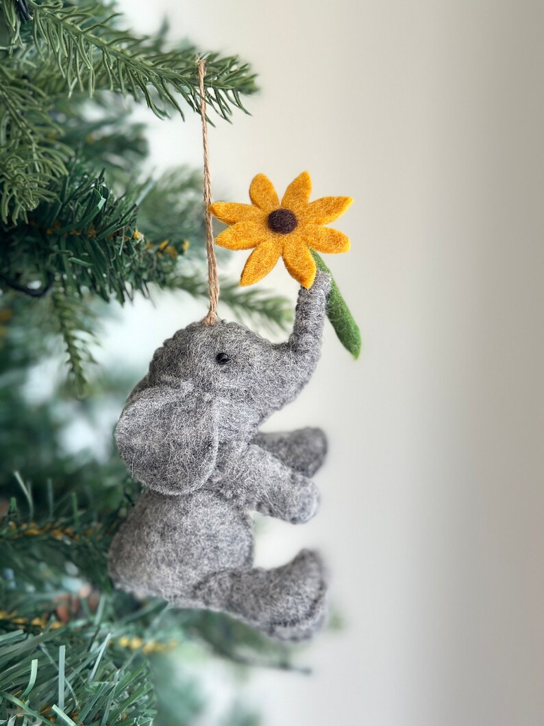 May include: A gray felt elephant ornament holding a yellow felt flower with a brown center. The ornament is hanging from a brown string on a green Christmas tree.