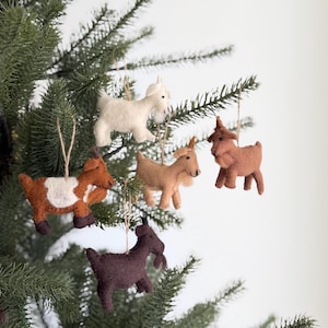 May include: Five felt goat ornaments in various colours including white, light brown, dark brown, and a brown and white spotted pattern. Each goat figure features a small black eye and is suspended by a thin tan string for hanging on a tree branch.