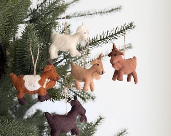 May include: Five felt goat ornaments in various colors including white, light brown, dark brown, and a brown and white spotted pattern. Each goat figure features a small black eye and is suspended by a thin tan string for hanging on a tree branch.
