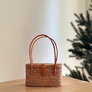 May include: A rectangular, woven rattan handbag with a leather strap and clasp. The bag is a light brown color and has two arched handles. The clasp is secured with a round, brown button. The bag is sitting on a wooden surface.