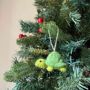 May include: A green felt turtle ornament hanging on a Christmas tree. The turtle has a brown eye and a string for hanging.
