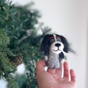 May include: A black and white felt dog ornament with a red nose and brown eyes. The ornament is being held in a person's hand.