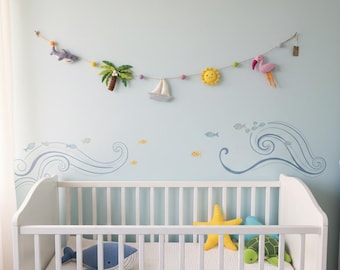 Könnte beinhalten: Eine Filzgirlande mit Hai, Palme, Segelboot, Sonne und Flamingo über einem weißen Kinderbett. Im Bett liegen ein blauer Wal, ein gelber Seestern und eine grüne Schildkröte als Plüschtiere. An der Wand befinden sich blaue Wellenaufkleber.