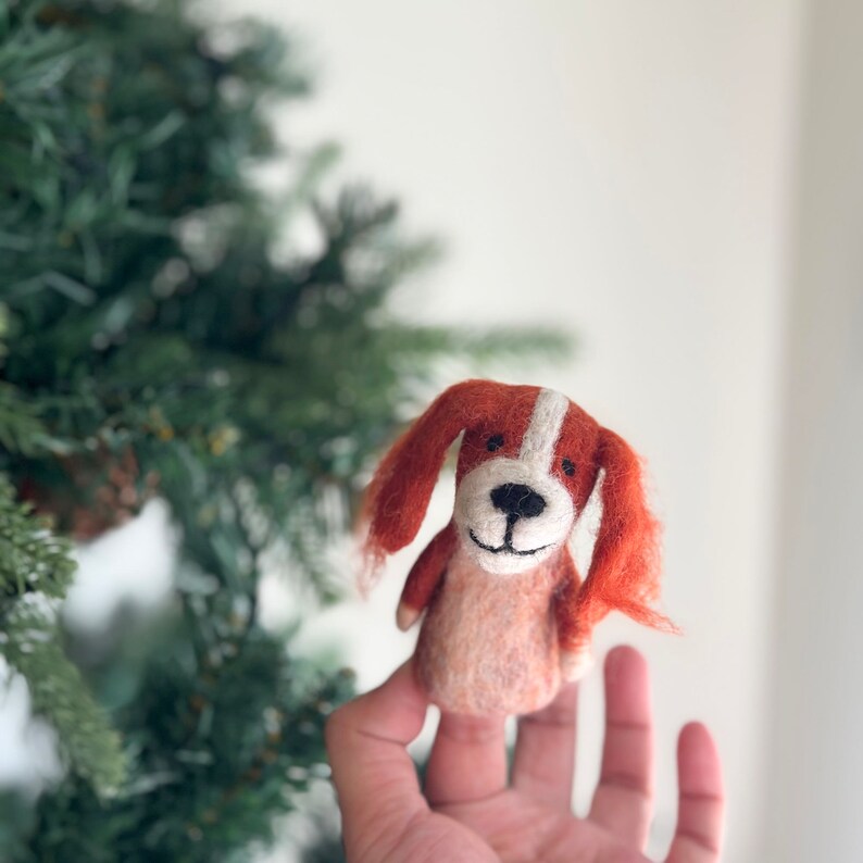 May include: A finger puppet of a dog with orange fur, white markings, and a black nose. The puppet is made of felt and has a smiling face.