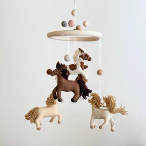 May include: A baby mobile featuring four felt horses in shades of brown and cream, suspended from a white ring with hanging felt balls. The horses have detailed manes and tails, creating a whimsical nursery decoration.