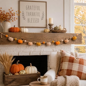 May include: A wooden mantelpiece decorated with a felt pumpkin garland, various small pumpkins, a pillar candle, and a framed sign reading GRATEFUL THANKFUL BLESSED. Below, a wooden crate holds a large orange pumpkin, a white pumpkin, and wheat stalks.