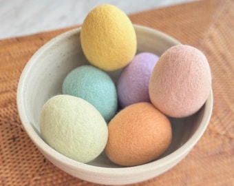May include: A ceramic bowl filled with six felt Easter eggs in pastel colors: yellow, light green, blue, purple, peach, and orange. The eggs are nestled in a white ceramic bowl, creating a soft and inviting display.