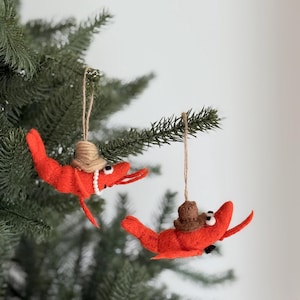 May include: Two red shrimp Christmas ornaments hanging from a green tree. Each shrimp wears a brown hat and has a string for hanging. One shrimp has a pearl necklace. The ornaments are made of felt.