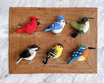 May include: A collection of six felt bird figurines in various colors, including red, blue, yellow, and green. The birds are arranged on a woven brown mat, showcasing detailed craftsmanship and vibrant hues. The birds are a cardinal, blue jay, hummingbird, chickadee, goldfinch, and a kingfisher.