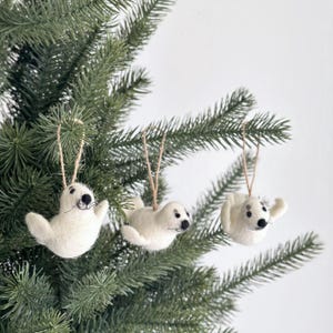 Pode incluir: Três enfeites de foca de feltro branco pendurados em uma árvore de Natal verde. As focas têm olhos e narizes pretos e estão pendurados em um barbante.