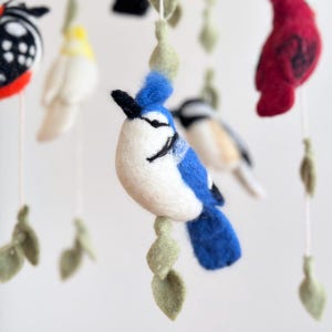 Puede incluir: Un m&oacute;vil de fieltro para beb&eacute; con un arrendajo azul, un cardenal, una mariposa y otras aves. El m&oacute;vil est&aacute; hecho de fieltro y tiene una base de madera natural.