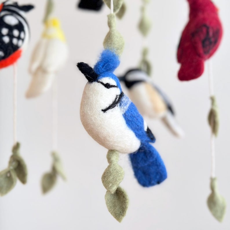 Puede incluir: Un m&oacute;vil de fieltro para beb&eacute; con un arrendajo azul, un cardenal, una mariposa y otras aves. El m&oacute;vil est&aacute; hecho de fieltro y tiene una base de madera natural.