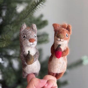 May include: Two felted squirrel figurines. One is gray with a brown acorn, the other is brown with a red acorn. Both have white eyes and are smiling. The background is a blurred green.