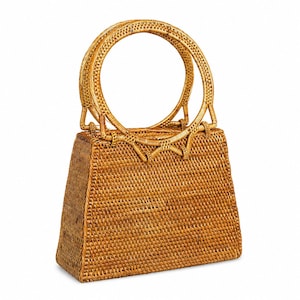 Può includere: Una borsa strutturata realizzata in rattan intrecciato color marrone chiaro. La borsa presenta un corpo trapezoidale con base piatta e due manici rigidi e circolari fissati sulla parte superiore. Il motivo dell'intreccio è uniforme.