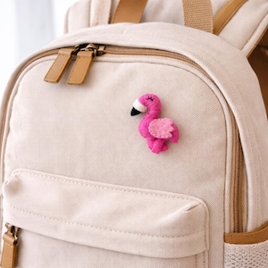 Puede incluir: Un broche de fieltro rosa con forma de flamenco, con pico blanco y negro y una pequeña ala rosa, está sujeto a una mochila beige. La mochila tiene detalles y cremalleras de cuero marrón.