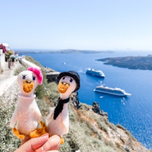 May include: Two handmade white felt goose dolls, one with a pink beret and pearl necklace, the other with a black hat and scarf. They have orange beaks and feet. The background features a blue sea, sky, and cruise ships.