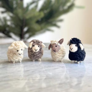 May include: Four felt sheep ornaments in white, brown, white and black. The sheep are standing on a white marble surface.