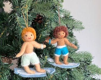 May include: Two felt ornaments of people surfing on blue felt surfboards. The person on the left has blonde hair and is wearing green swim trunks. The person on the right has brown hair and is wearing blue swim trunks.