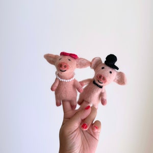 May include: Two pink felt pig finger puppets. One pig wears a red bow and pearl necklace, the other a black top hat and bow tie. The puppets are held up by a hand with red nail polish.