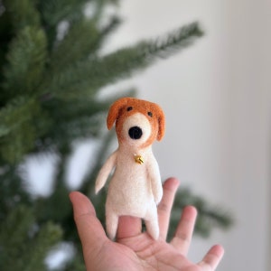 May include: A small, white felt dog with brown ears and a gold bell around its neck. The dog is sitting on a person's finger.