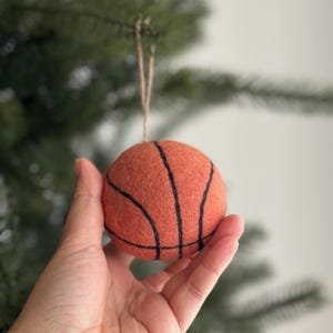 Puede incluir: Adorno de baloncesto de fieltro con líneas negras, suspendido por una cuerda de hilo natural. La esfera naranja se sostiene en una mano, con un fondo verde borroso. Esta decoración de temática deportiva es adecuada para un árbol de Navidad.