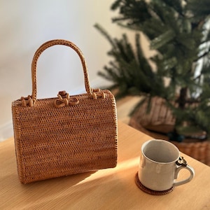 May include: A woven rattan handbag with a curved handle and a bow-shaped clasp. The bag is a warm brown color and sits on a light-colored wooden surface. A ceramic mug with a decorative handle and a coaster are also on the table.