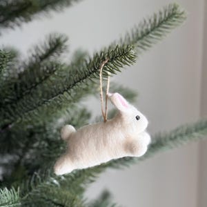 May include: A white felt bunny ornament with pink inner ears and black eyes hangs from a natural twine string. The bunny is suspended from a green Christmas tree branch, creating a festive holiday decoration.