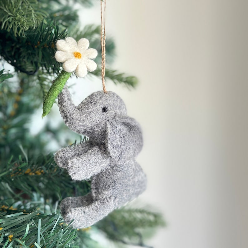 May include: A gray felt elephant ornament holding a white felt flower with a yellow center. The ornament is hanging from a tree branch.