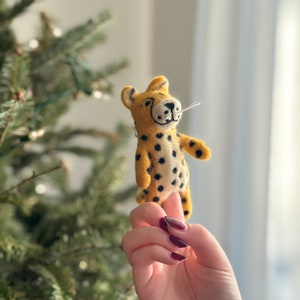 May include: A yellow and black felt cheetah ornament with a white felt nose and whiskers. The ornament is being held by a hand with dark red nail polish.