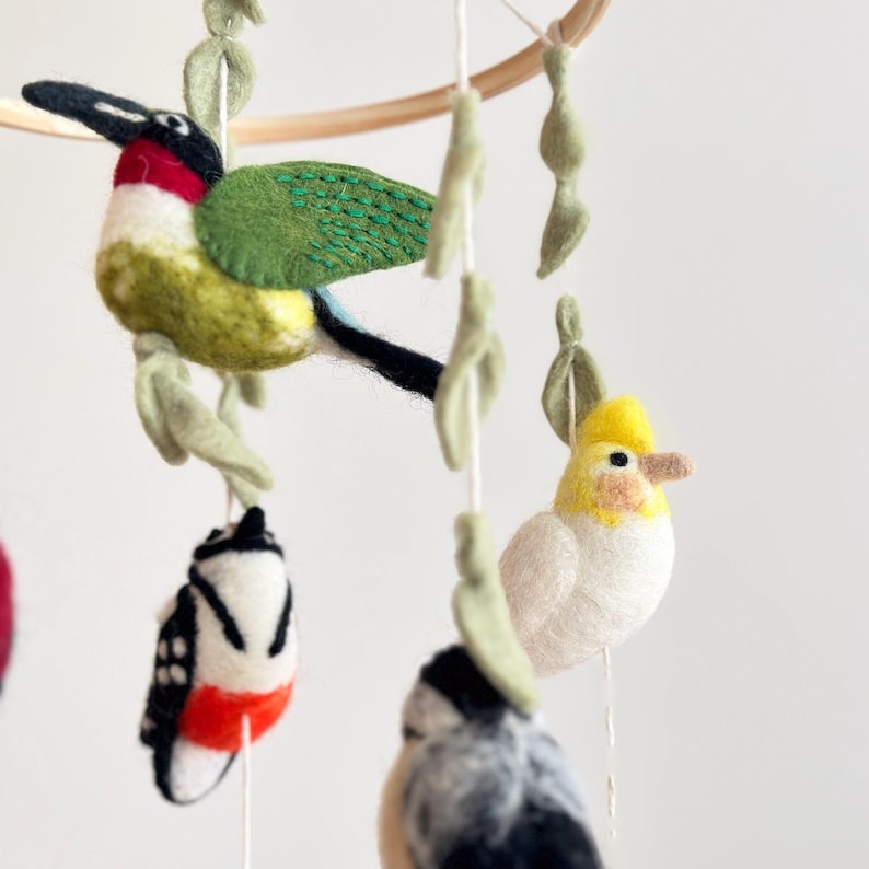 Puede incluir: Un m&oacute;vil de p&aacute;jaros de fieltro con p&aacute;jaros verdes, amarillos, blancos, rojos y negros colgando de un aro de madera. Los p&aacute;jaros est&aacute;n suspendidos de un hilo blanco con hojas de fieltro verde.