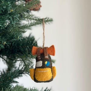 Puede incluir: Un adorno de fieltro con forma de mochila verde y amarilla con correas naranjas. La mochila tiene un pequeño parche azul y amarillo en la parte delantera.