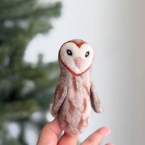 Op de afbeelding: Een handgemaakt vilten uilbeeldje. De uil heeft een witte kop met een hartvormige omtrek, zwarte ogen en een roze snavel. Het lichaam is lichtbruin en beige met donkerbruine markeringen. De uil is ongeveer 10 cm hoog.