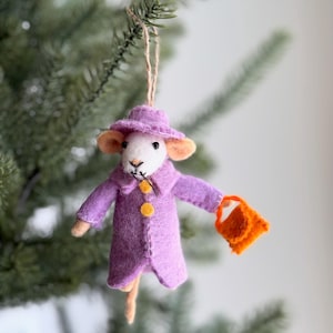 May include: A whimsical felt Christmas ornament of a mouse wearing a purple coat and hat, holding an orange purse. The mouse has tan ears and a long tail, and is suspended by a twine string.