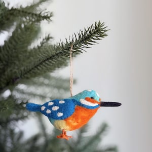 May include: A felt bird ornament with a blue head and wings, orange chest, and white polka dots. The bird has a black beak and is hanging from a natural twine string. The ornament is displayed on a green Christmas tree branch.