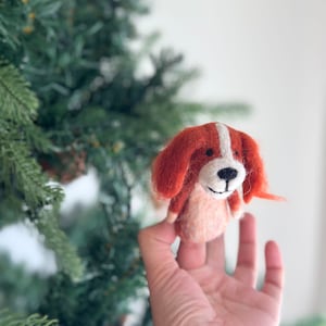 May include: A felt finger puppet of a dog with orange and white fur. The dog has a white nose and black eyes.