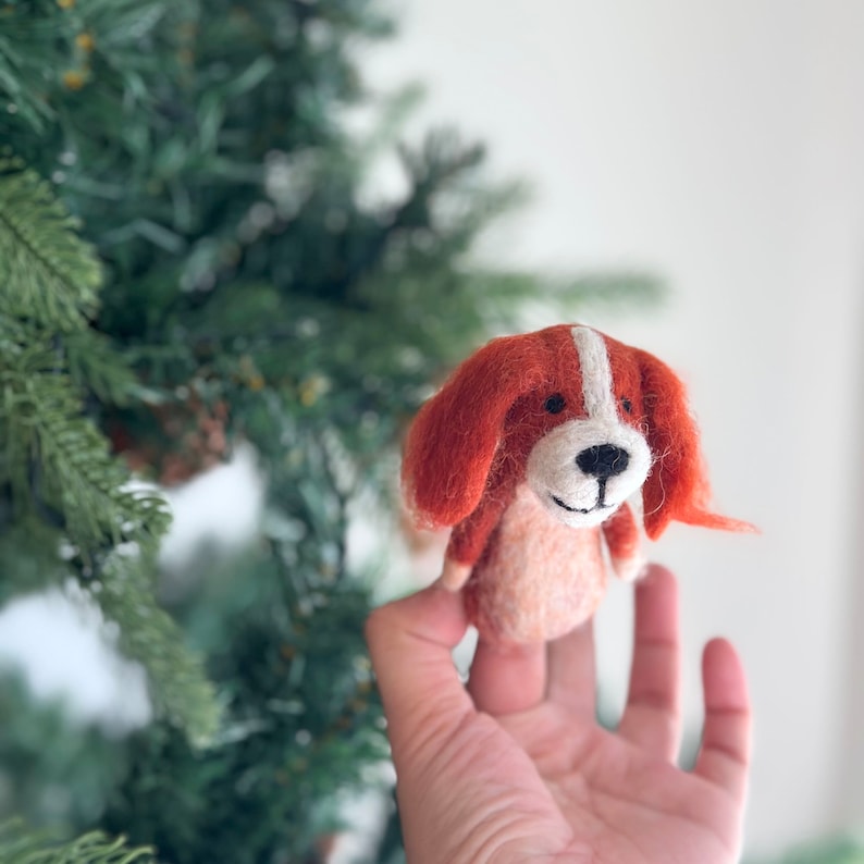 May include: A felt finger puppet of a dog with orange and white fur. The dog has a white nose and black eyes.